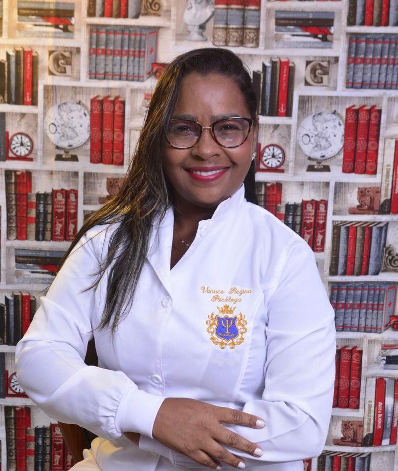 Mulher sorridente de cabelos castanhos escuros, usando blusa clara, sentada em ambiente iluminado e acolhedor, representando a psicóloga Vanuce Regina